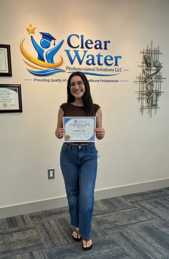 Clear Water graduate smiling with a certificate of completion at the studio logo wall